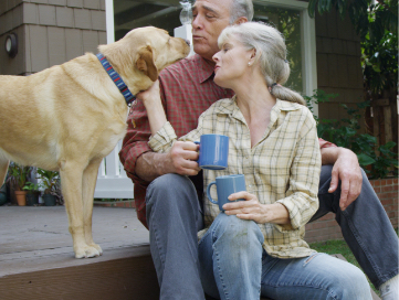Older man and woman sitting on front porch steps with enjoying morning coffee while petting their golden lab. 