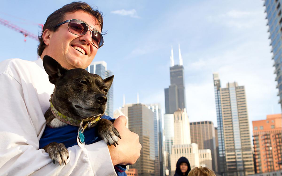 Man in black sunglasses holding small black dog on a boat with Chicago skyline in background
