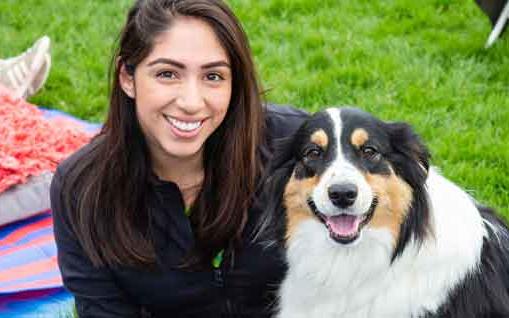 Woman with long brown hair and black fleece jacket on sits next to black, tan, and white fluffy dog