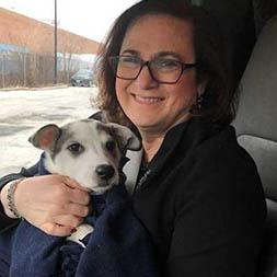 A woman with brown hair and glasses sits in a car holding a white dog