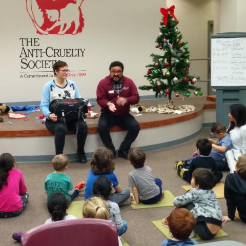 Woman in blue shirt sitting next to man in red sweatshirt next to christmas tree talking to children