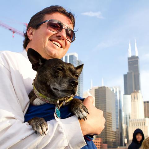 Man in black sunglasses on a boat holding small black dog with Chicago skyline in background