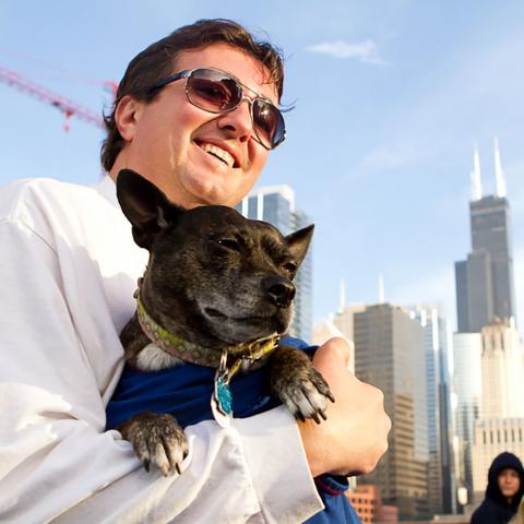White man with brown hair wearing sunglasses holding small dog with Chicago skyline in background